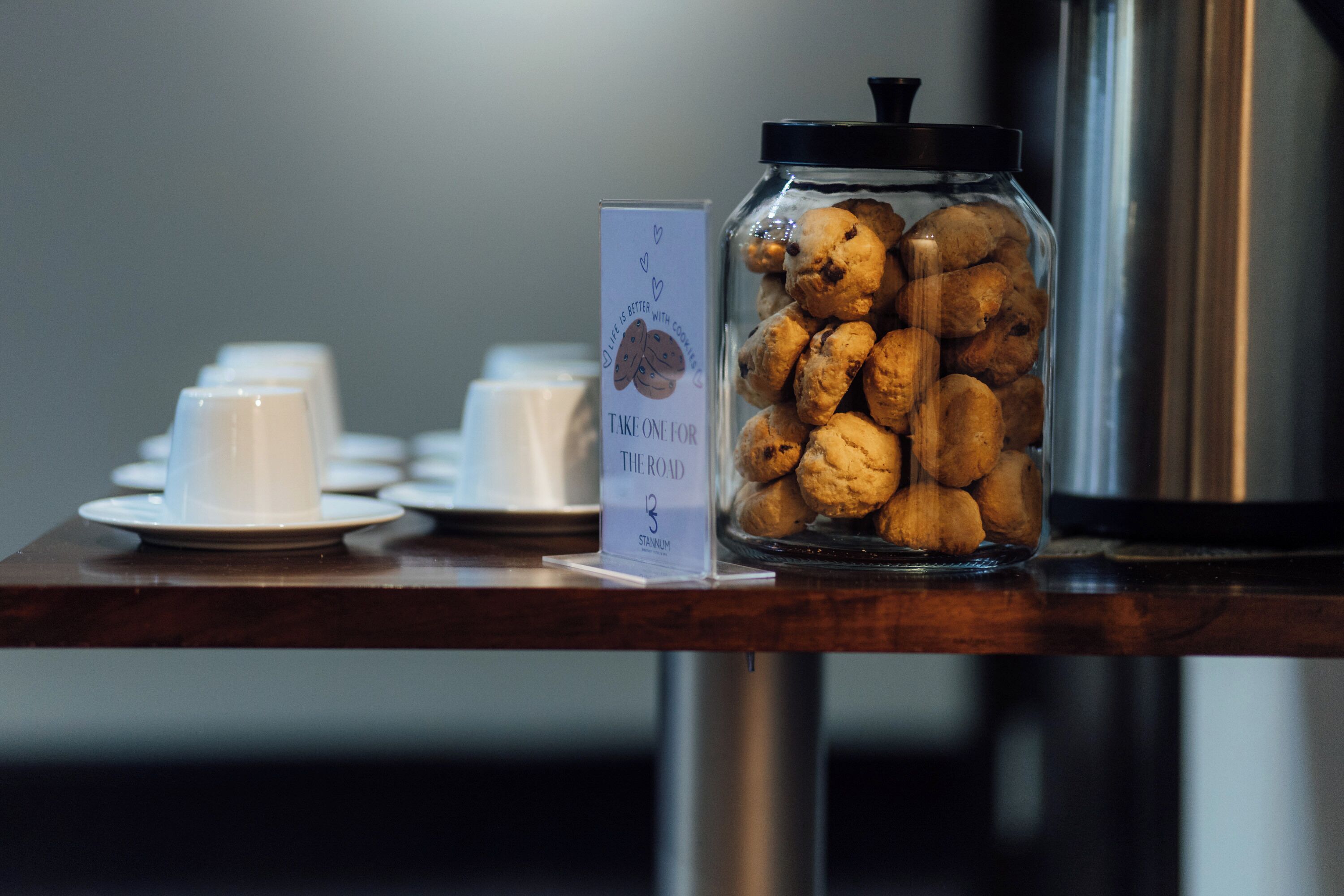 Un tarro de cristal lleno de galletas sobre una mesa de madera, junto con un cartel y tazas blancas.