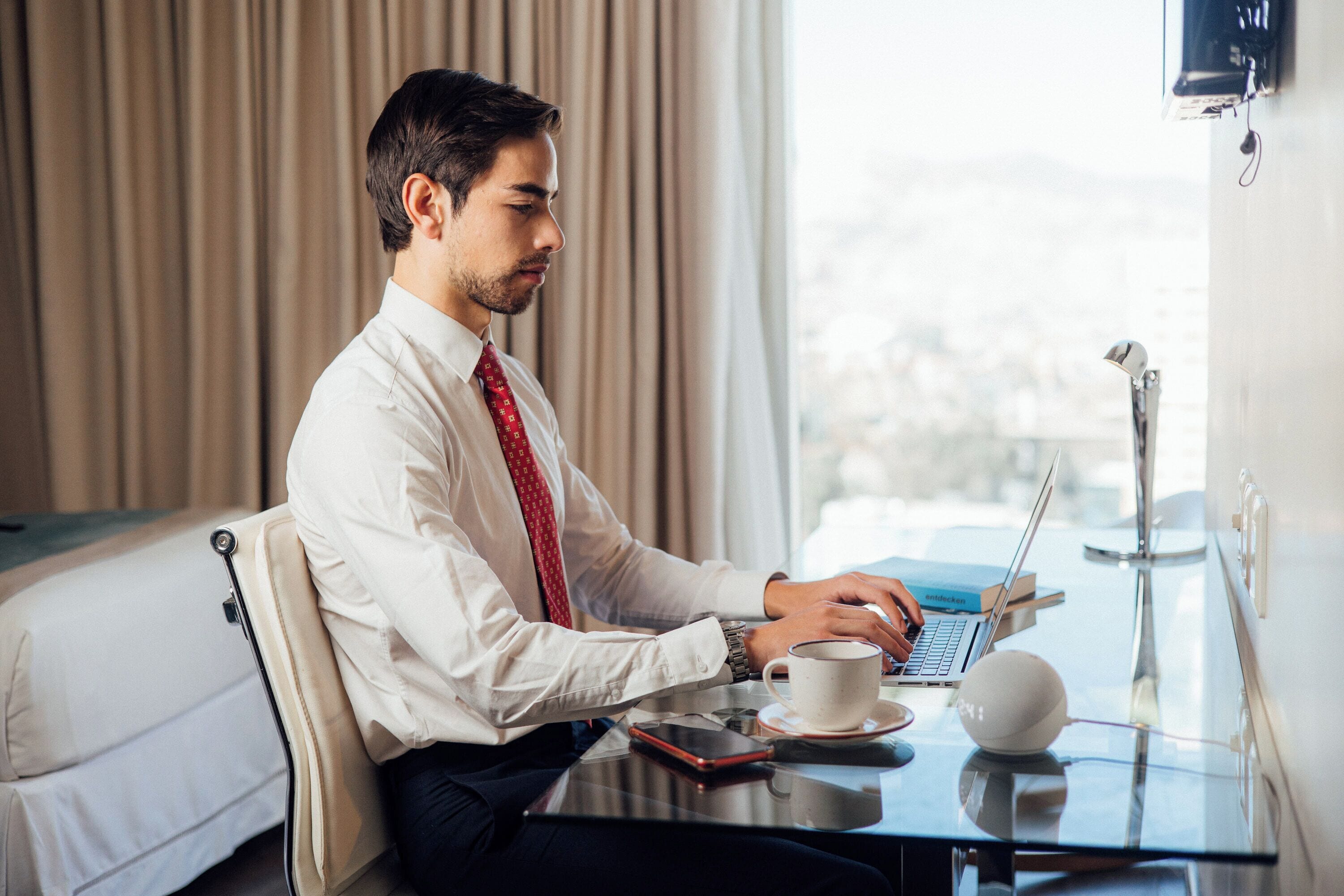 A man in a white shirt and red tie working on a laptop in a hotel room with a bed, a cup, and a remote control on the table.