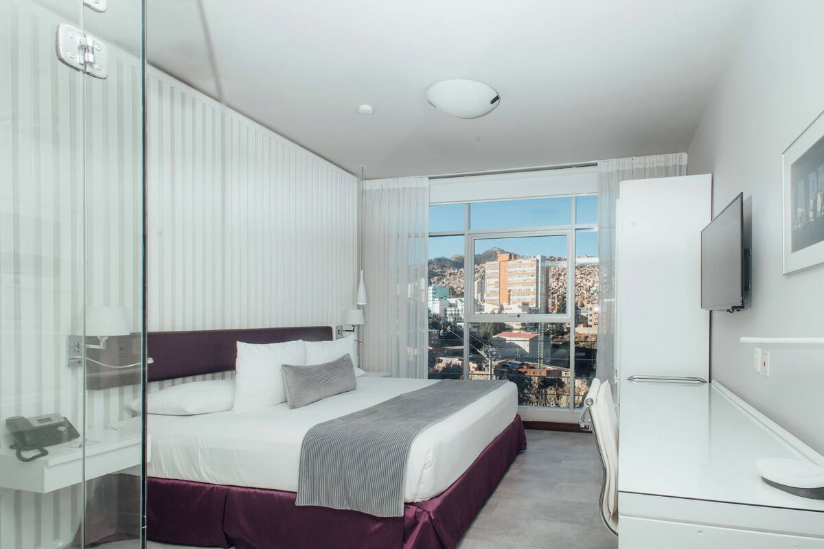 Habitación de hotel moderna con una cama grande, un escritorio, un televisor y vistas a la ciudad desde la ventana.