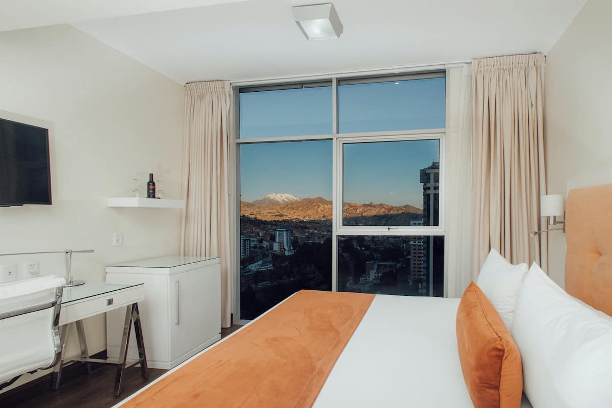 Habitación de hotel con un gran ventanal, una cama con ropa de cama blanca y naranja, un escritorio con una silla, un televisor de pantalla plana montado en la pared y un estante pequeño con una botella y vasos.