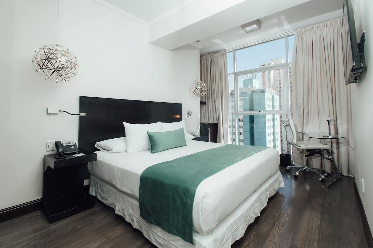 Una habitación de hotel moderna con una cama grande, una mesita de noche con teléfono, una silla y vistas de la ciudad a través de la ventana.