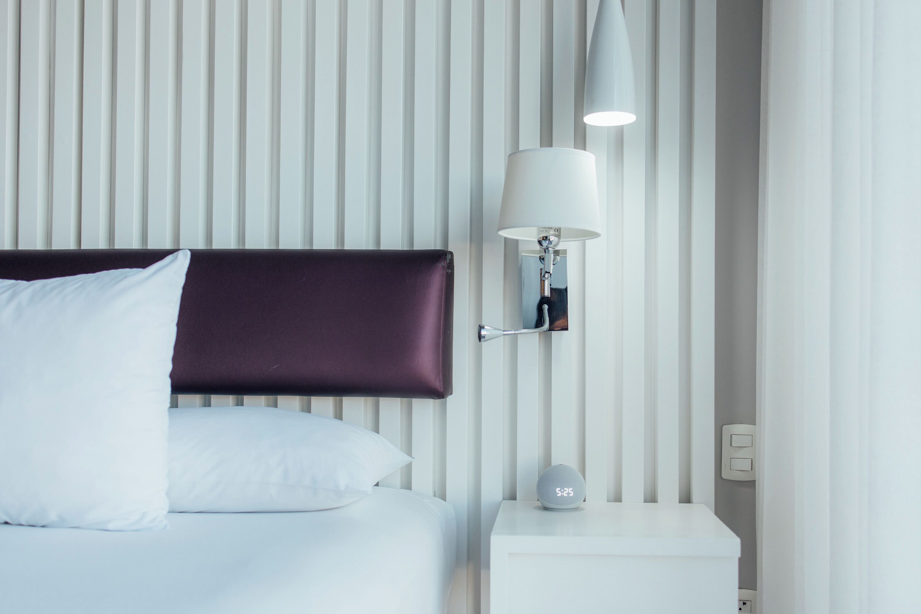A modern hotel room with a bed, white bedding, a dark headboard, a bedside table with a lamp, and a clock displaying 5:25.
