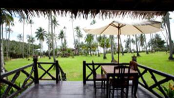 A covered outdoor area with a table and chairs, a thatched roof, and a view of palm trees.