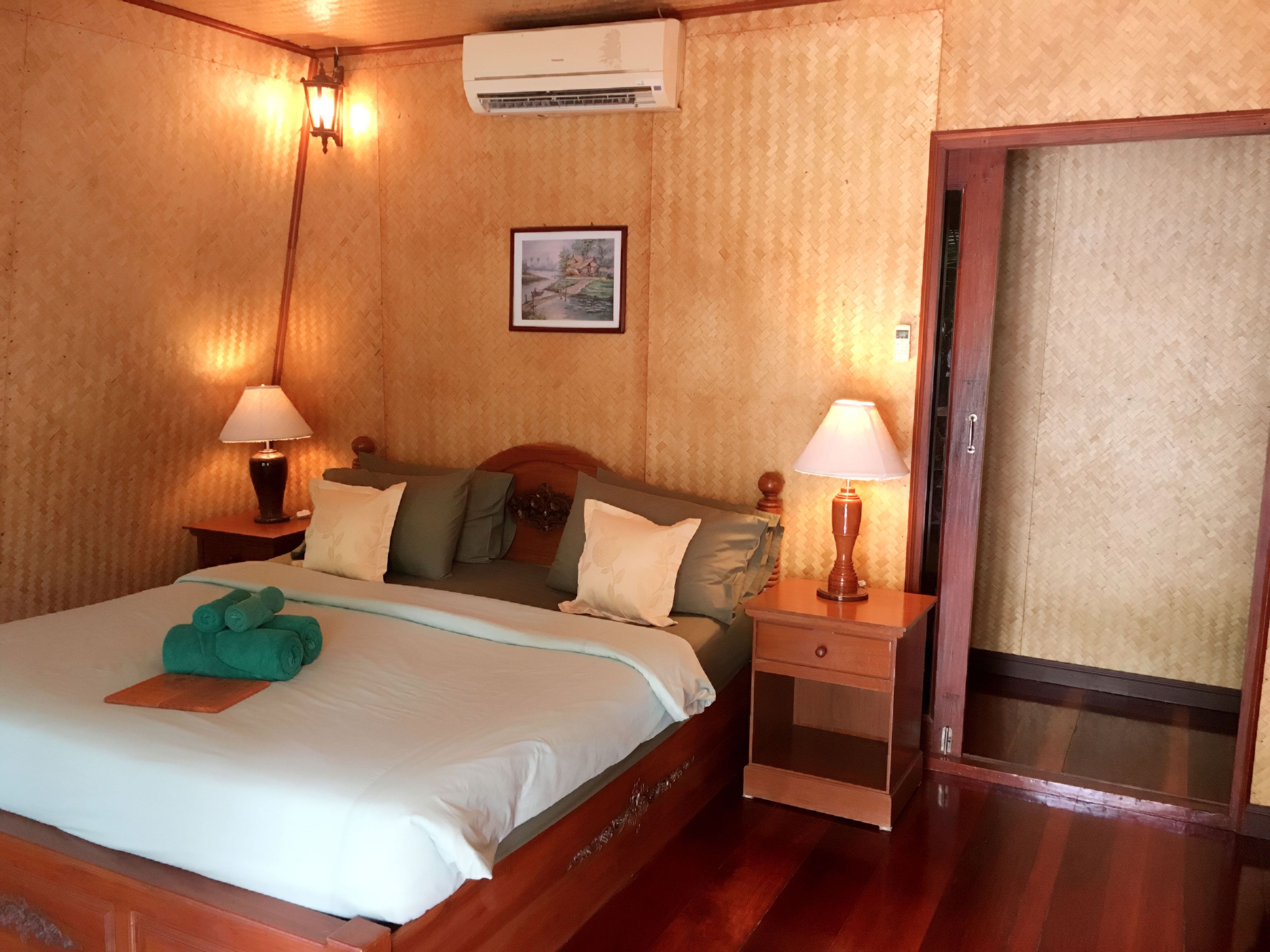A hotel room with a wooden bed, two bedside tables with lamps, a wall-mounted air conditioner, and a door leading to another room.
