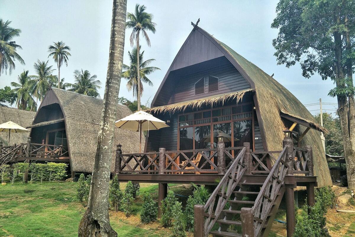 Un bungalow au toit de chaume, doté d’une terrasse et d’un escalier en bois, entouré de palmiers.