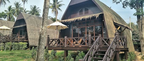 A thatched-roof bungalow with a wooden deck and staircase, surrounded by palm trees.