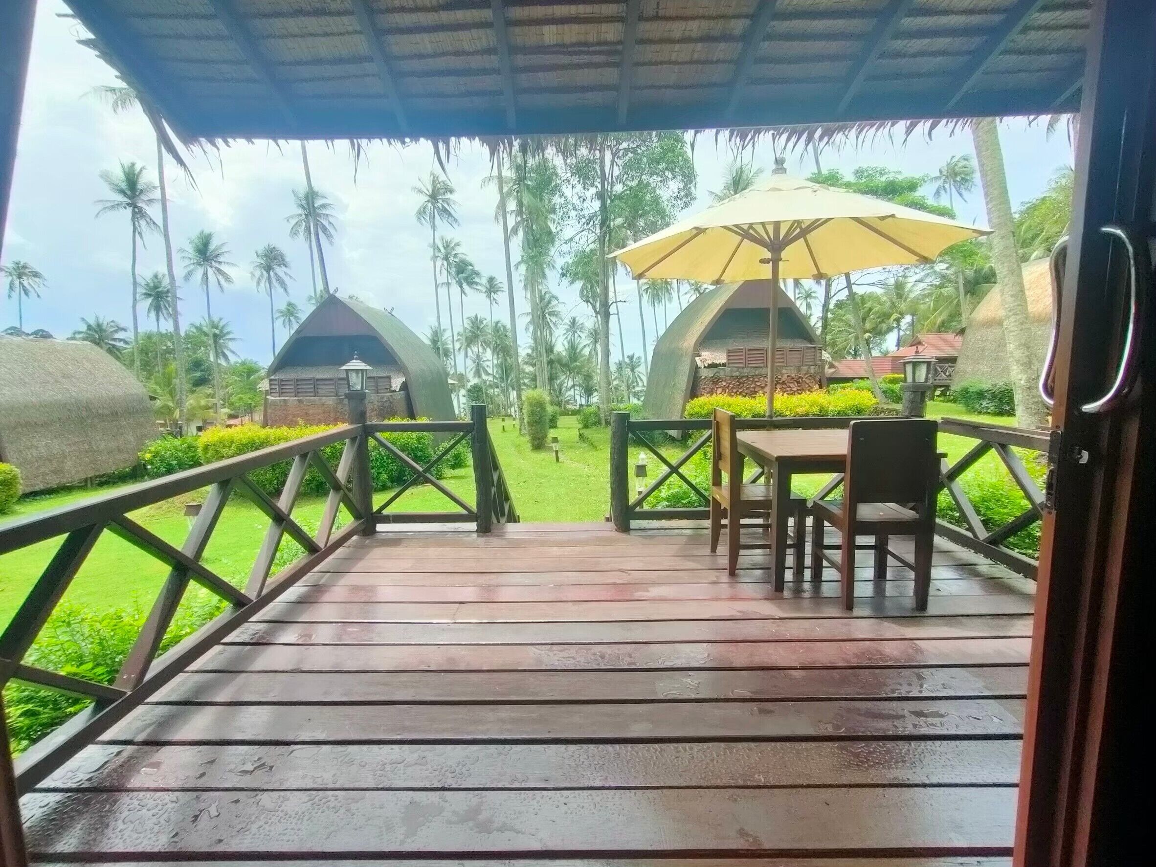 Una terraza de madera con una mesa y sillas, un paraguas amarillo y dos cabañas en forma de cúpula en un entorno tropical.