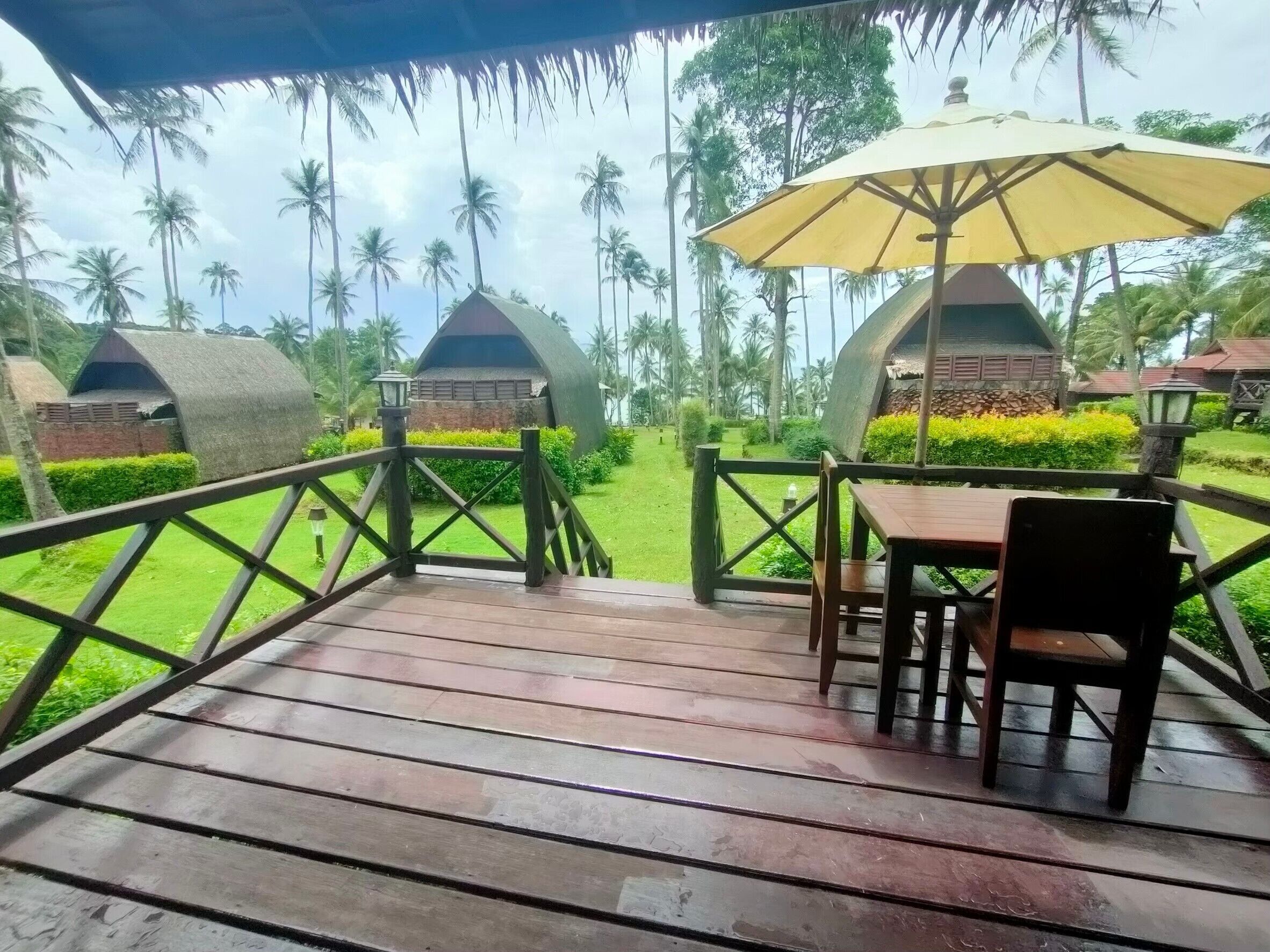 Um deck de madeira com mesa e cadeiras, um guarda-sol amarelo e vista para bangalôs sobre a água.