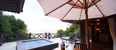 A hot tub, wooden table, and lounge chairs on a balcony with a view of the sea.