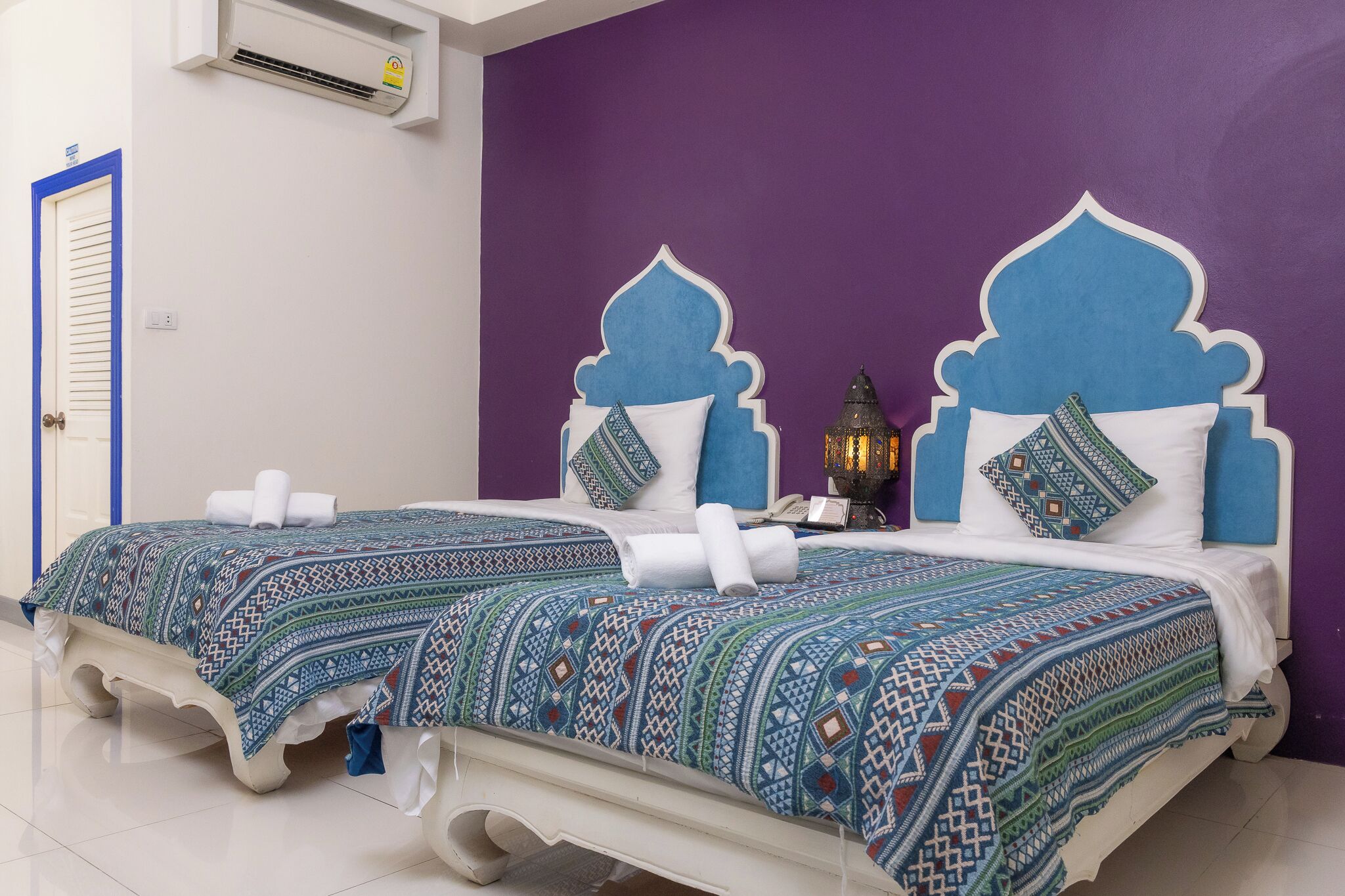 Una habitación con dos camas, cada una con colcha y almohadas estampadas, una mesita con lámpara y un aire acondicionado instalado en la pared.