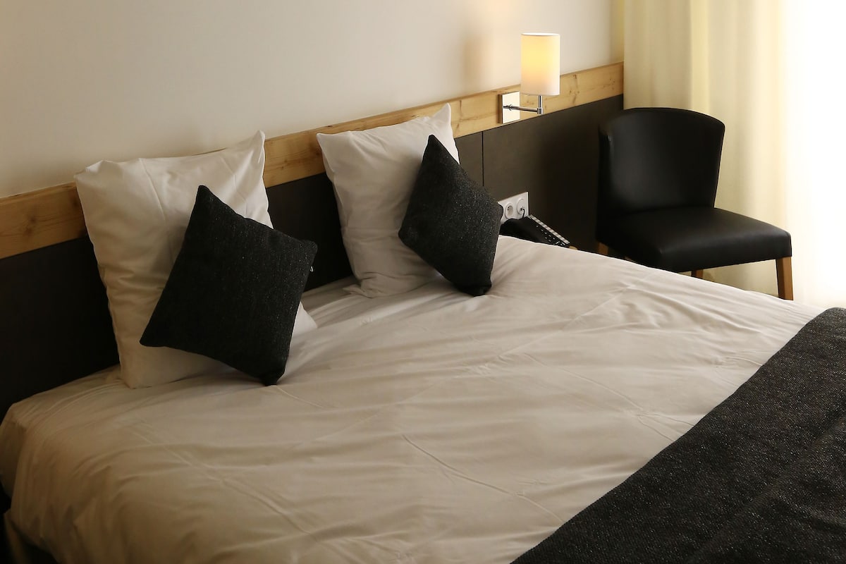 A hotel room with a bed, a wooden headboard, a wall-mounted light, a black chair, and a window with curtains.