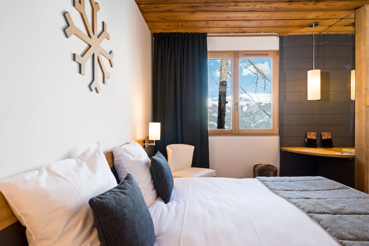 A bedroom with a large bed, a wooden ceiling, a window with a view of snow-capped mountains, a wall-mounted light fixture, and a small table with chairs.