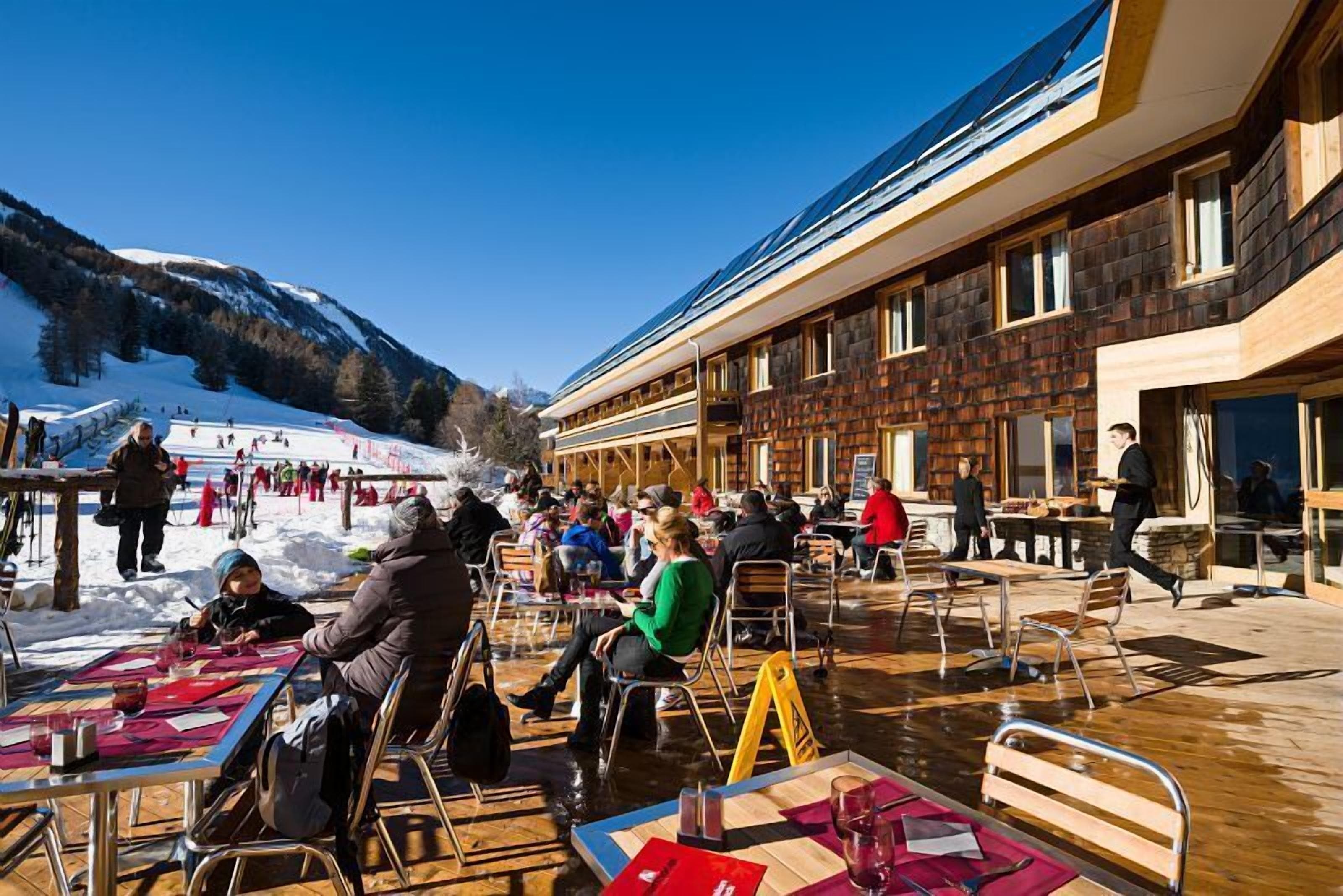 Un espacio al aire libre con mesas y sillas, un edificio de madera con ventanas amplias y una montaña nevada al fondo.