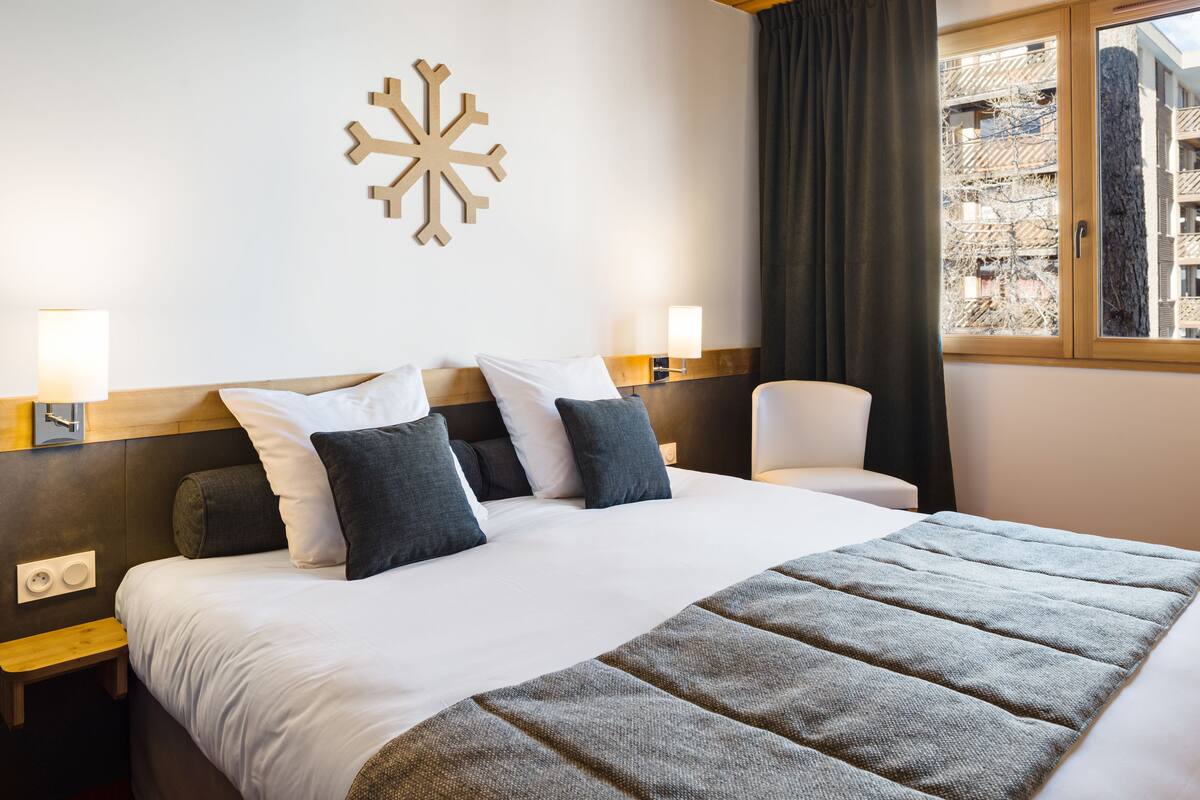 A hotel room with a large bed, two bedside tables, a chair, a window with a view of buildings, and a wall-mounted light fixture.