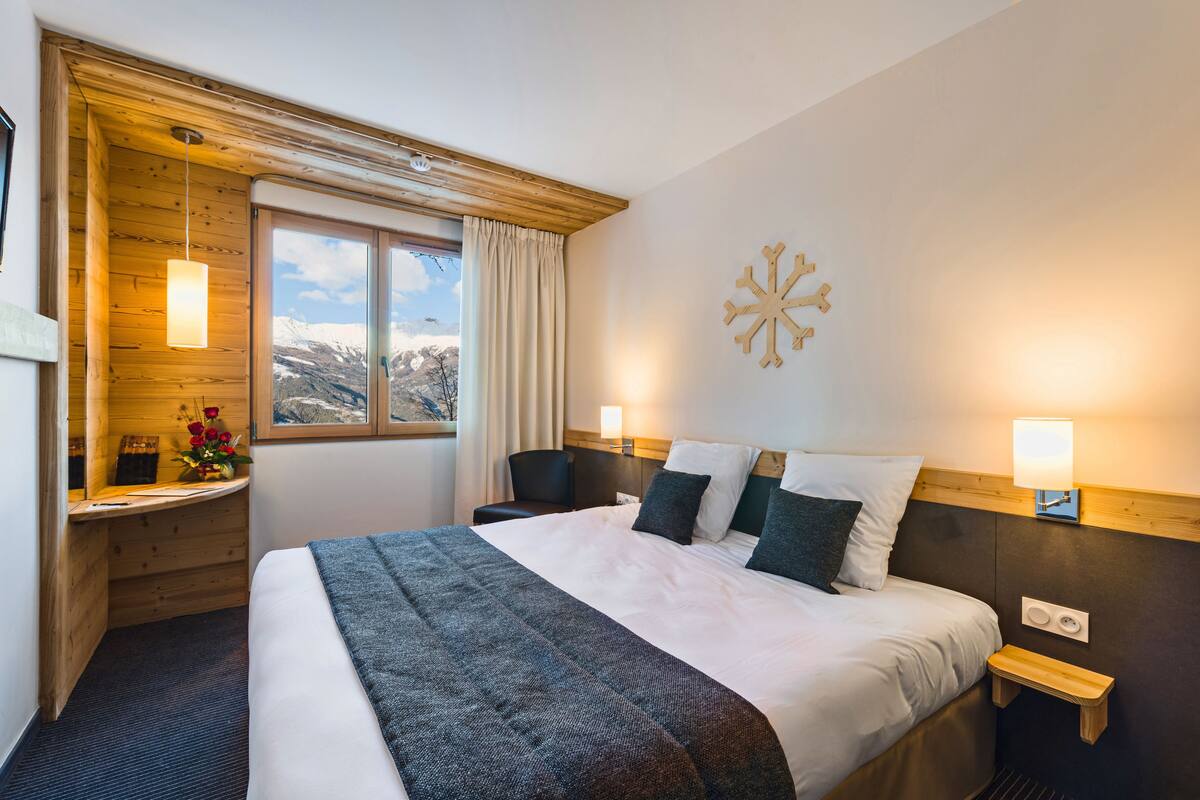 A hotel room with a large bed, wooden headboard, bedside tables with lamps, a view of mountains through a window, and a decorative wall piece.