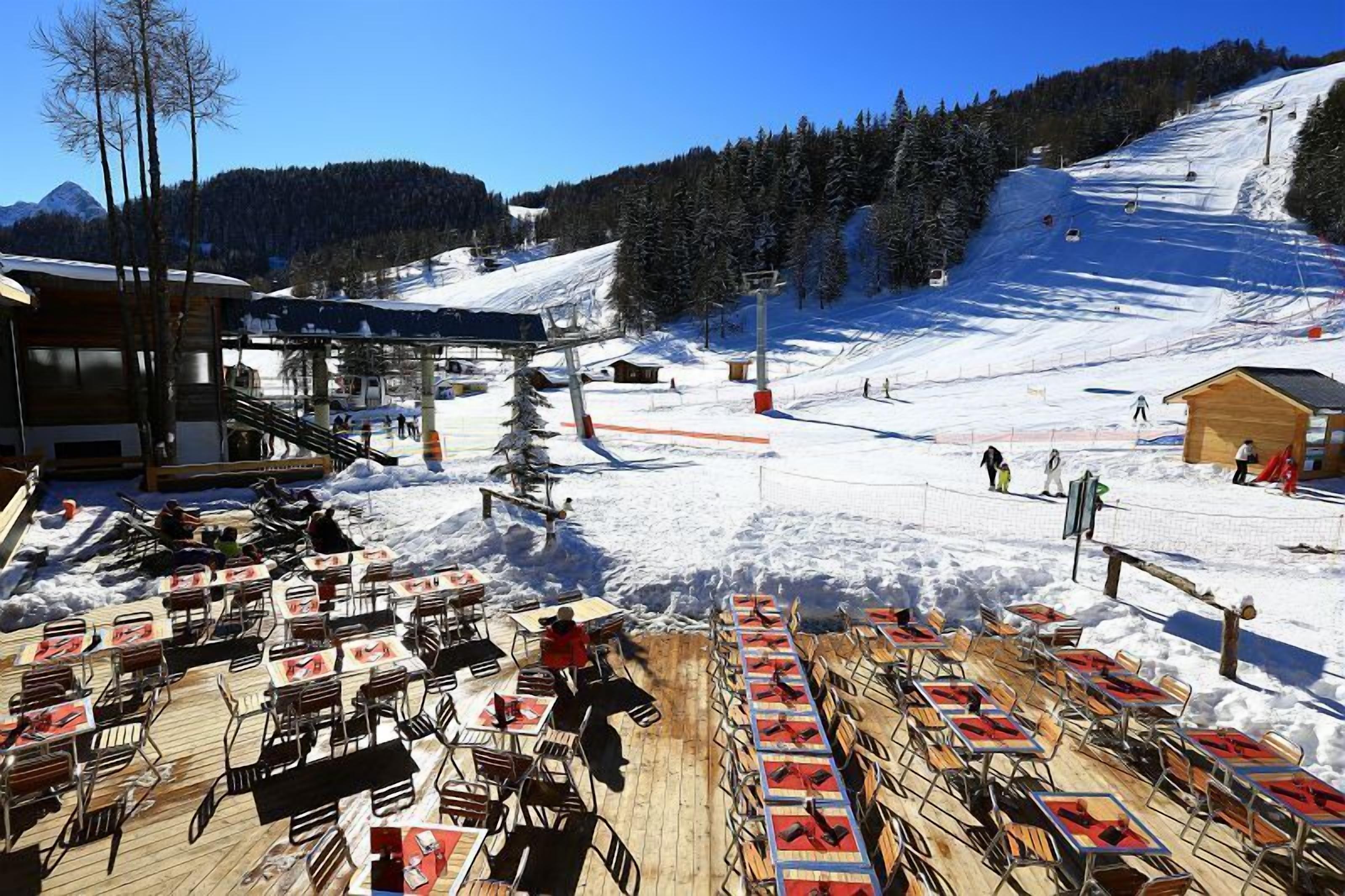 Un espace de restauration en plein air, couvert de neige, avec des tables et des chaises, un télésiège en arrière-plan et une vue sur les montagnes.