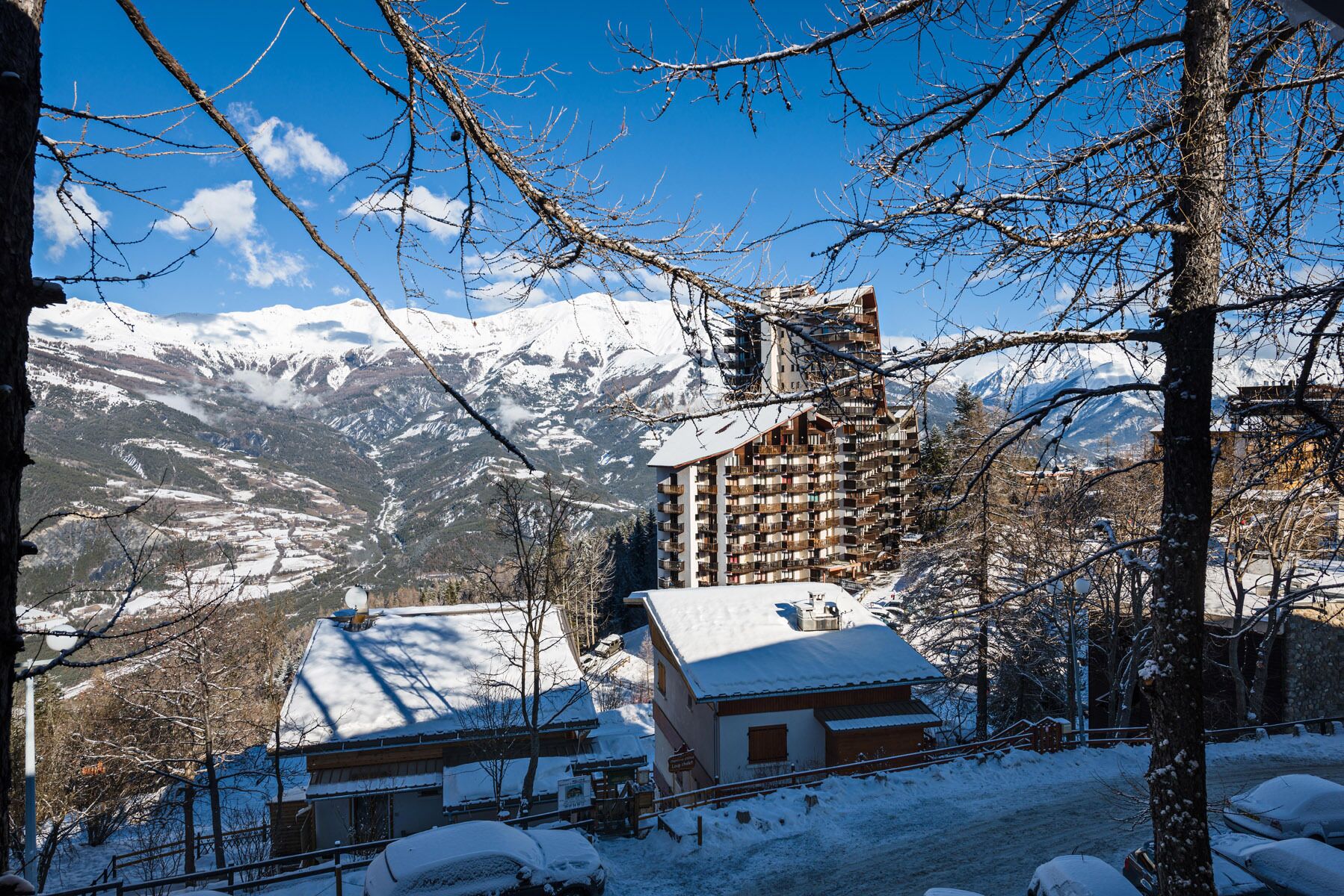 A mountain lodge with a snow-covered roof, a large building with balconies, and a view of snow-capped mountains.