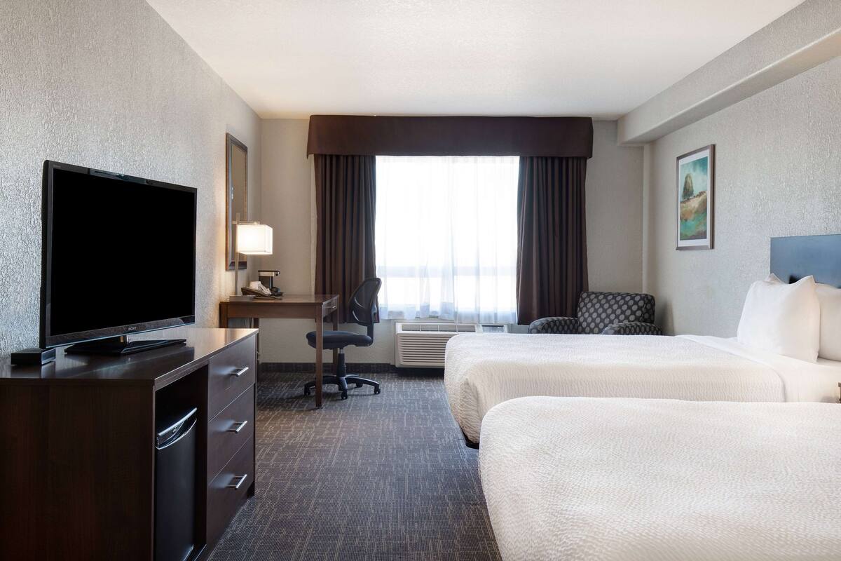 A hotel room with a flat-screen TV, a desk with a chair, two beds, a large window with curtains, and a painting on the wall.