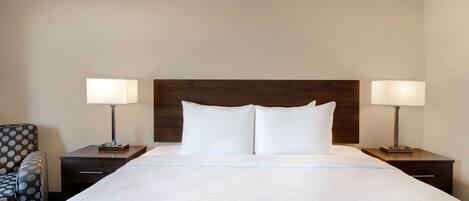 A hotel room with a large bed, two bedside tables with lamps, a patterned armchair, and a wooden headboard.