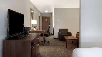 A hotel room with a flat-screen TV, a desk with a lamp, a chair, a sofa, and a bed.