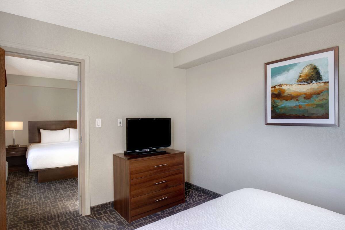 A hotel room with a bed, a wooden dresser with a flat-screen TV, a painting on the wall, and a carpeted floor.