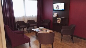 A room with a wooden floor, a table with magazines, four chairs, a TV on the wall, and a window with curtains.