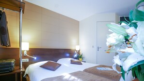 A hotel room with a bed, bedside lamps, a lamp on a table, a suitcase, a plant, and a door.