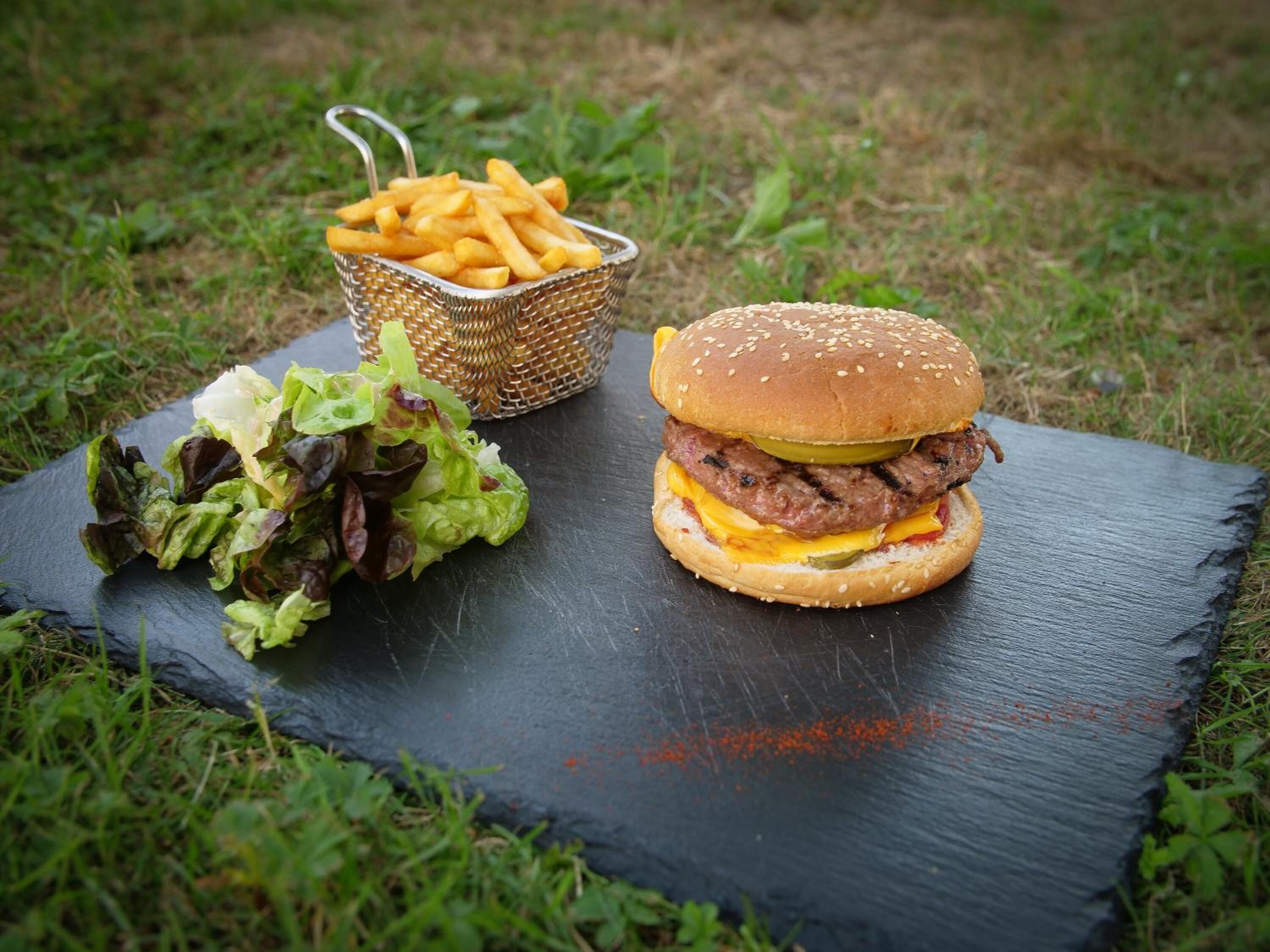 A burger with a sesame seed bun, cheese, and a pickle, served with a side of fries and a mixed green salad on a slate board.