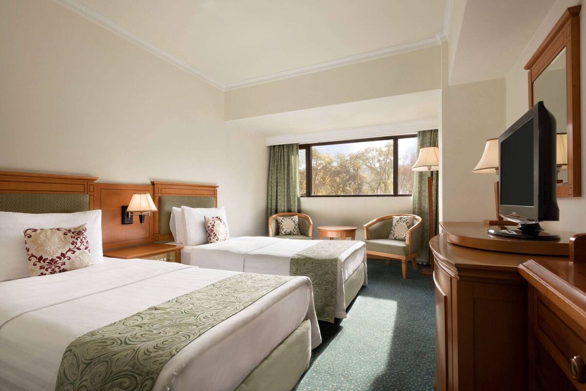 A hotel room with two beds, a television, a desk, and a window with a view of trees.