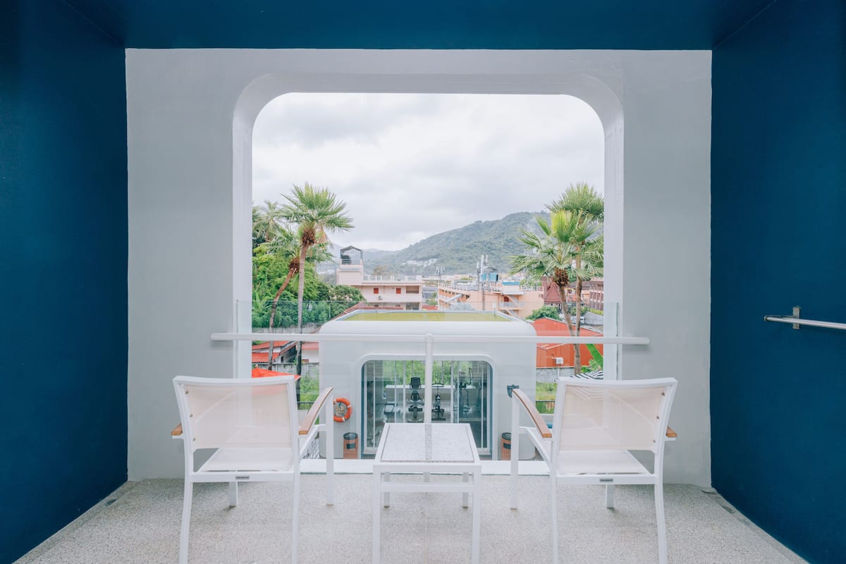Een balkon met twee witte stoelen en een tafeltje, met uitzicht op een stadsgezicht met gebouwen en palmbomen.