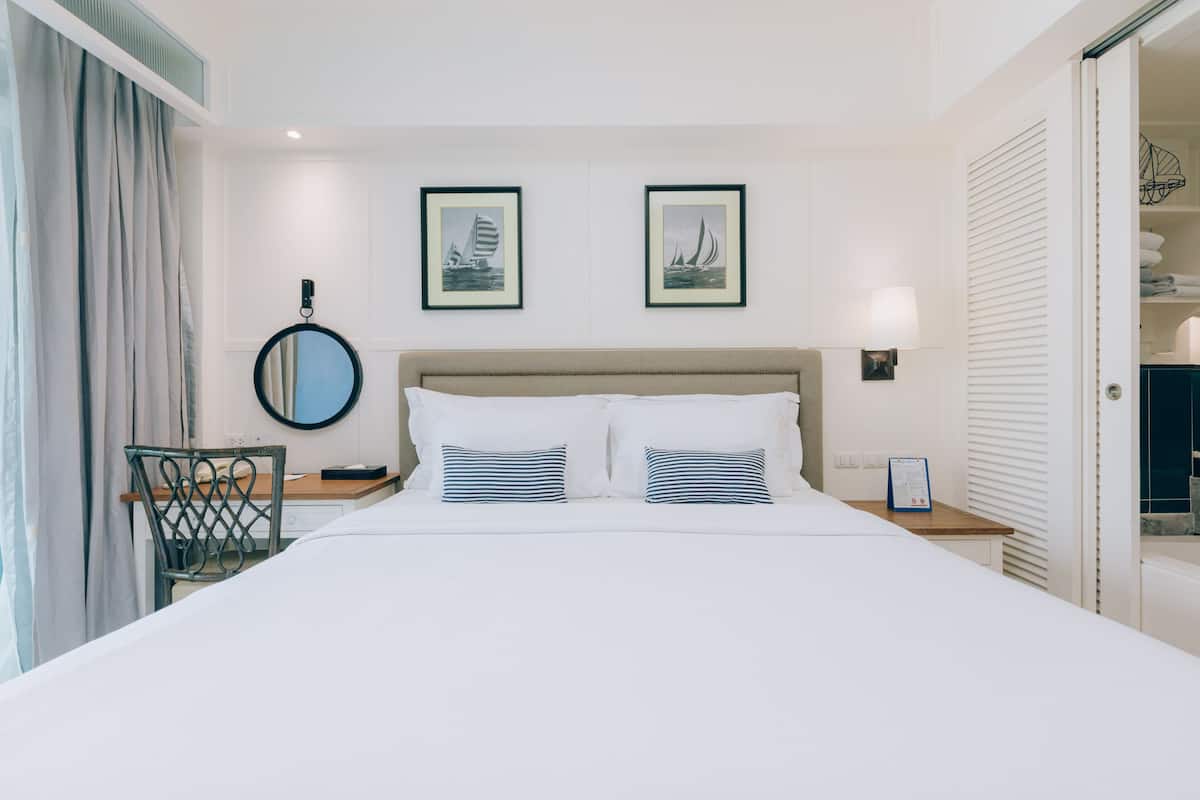 Een net opgemaakt bed met twee kussens, een nachtkastje met een lamp, een stoel, en twee ingelijste foto's aan de muur.