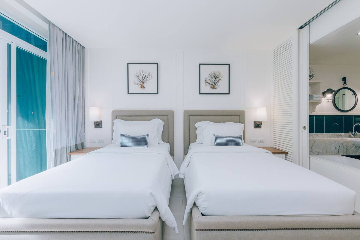 Een hotelkamer met twee bedden, een groot raam met gordijnen, een badkamer met een wastafel en spiegel, en wanddecoraties in lijsten.