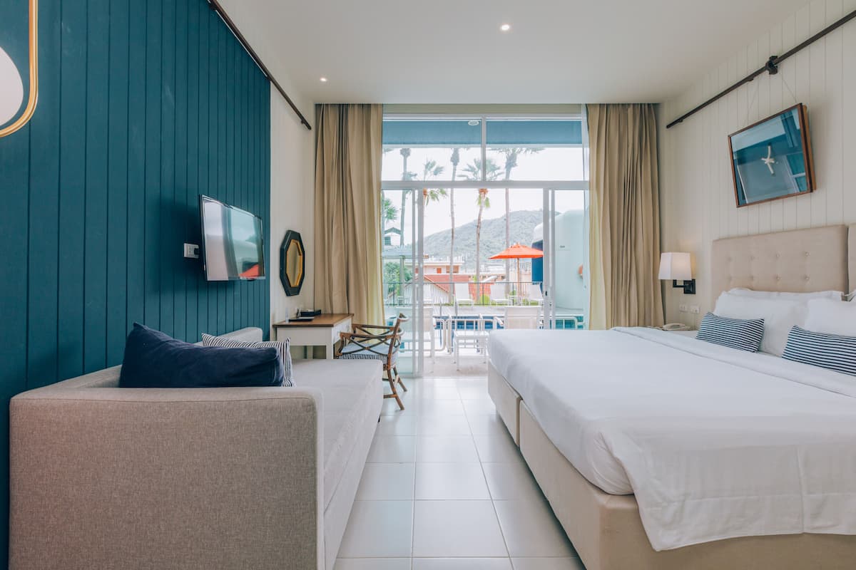 Een hotelkamer met een bed, een bank, een televisie, een bureau en uitzicht op het zwembad en palmbomen.