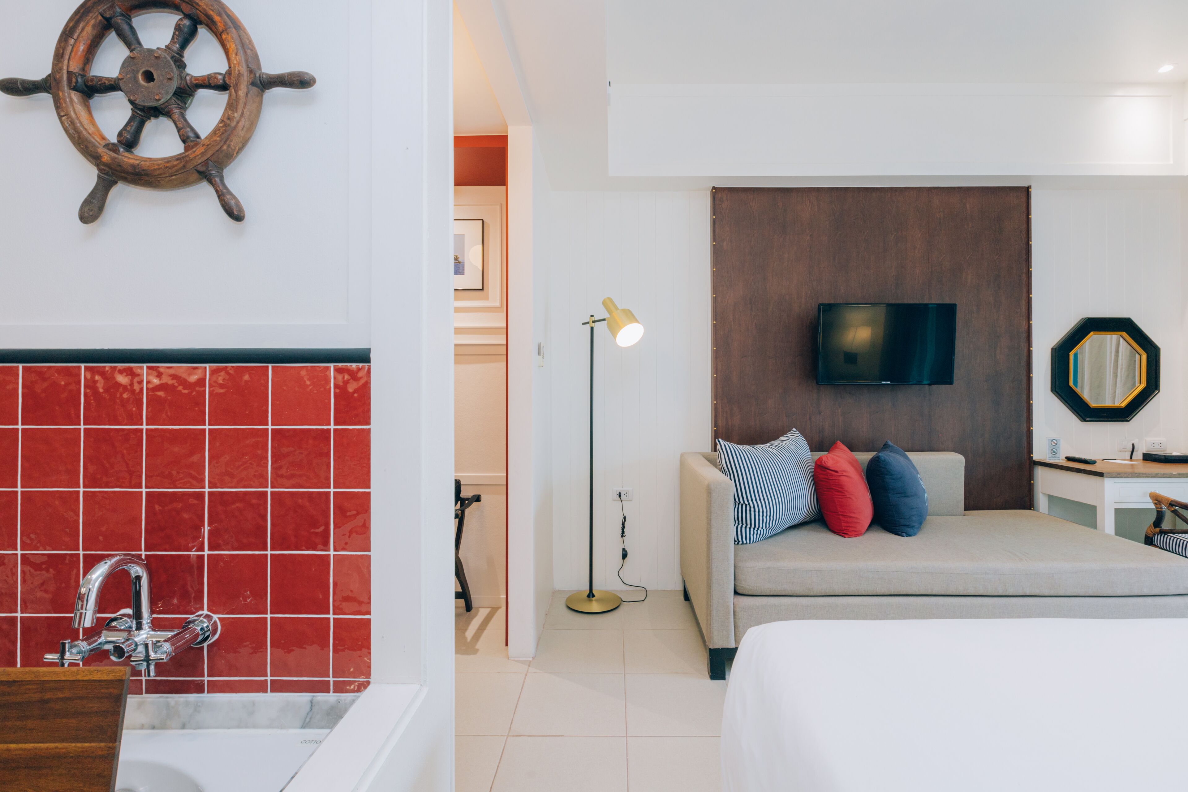 A modern hotel room with a red-tiled wall, a wooden ship's wheel decoration, a bed with multiple pillows, a bedside table with a lamp, and a television on the wall.