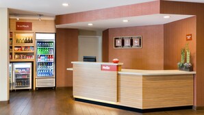 A reception area with a wooden reception desk, a 'Hello' sign, a 'Welcome' sign, a 'Stay' sign, a 'Meet' sign, and a 'Play' sign. There are two refrigerators with beverages and snacks, and a plant on the right side. - TownePlace Suites by Marriott Aberdeen (Aberdeen)