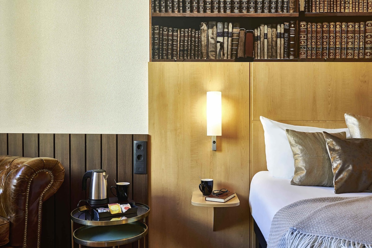 Habitación de hotel con cabecera de madera, mesita de noche con lámpara, cafetera y una pequeña mesita con un libro y vasos.