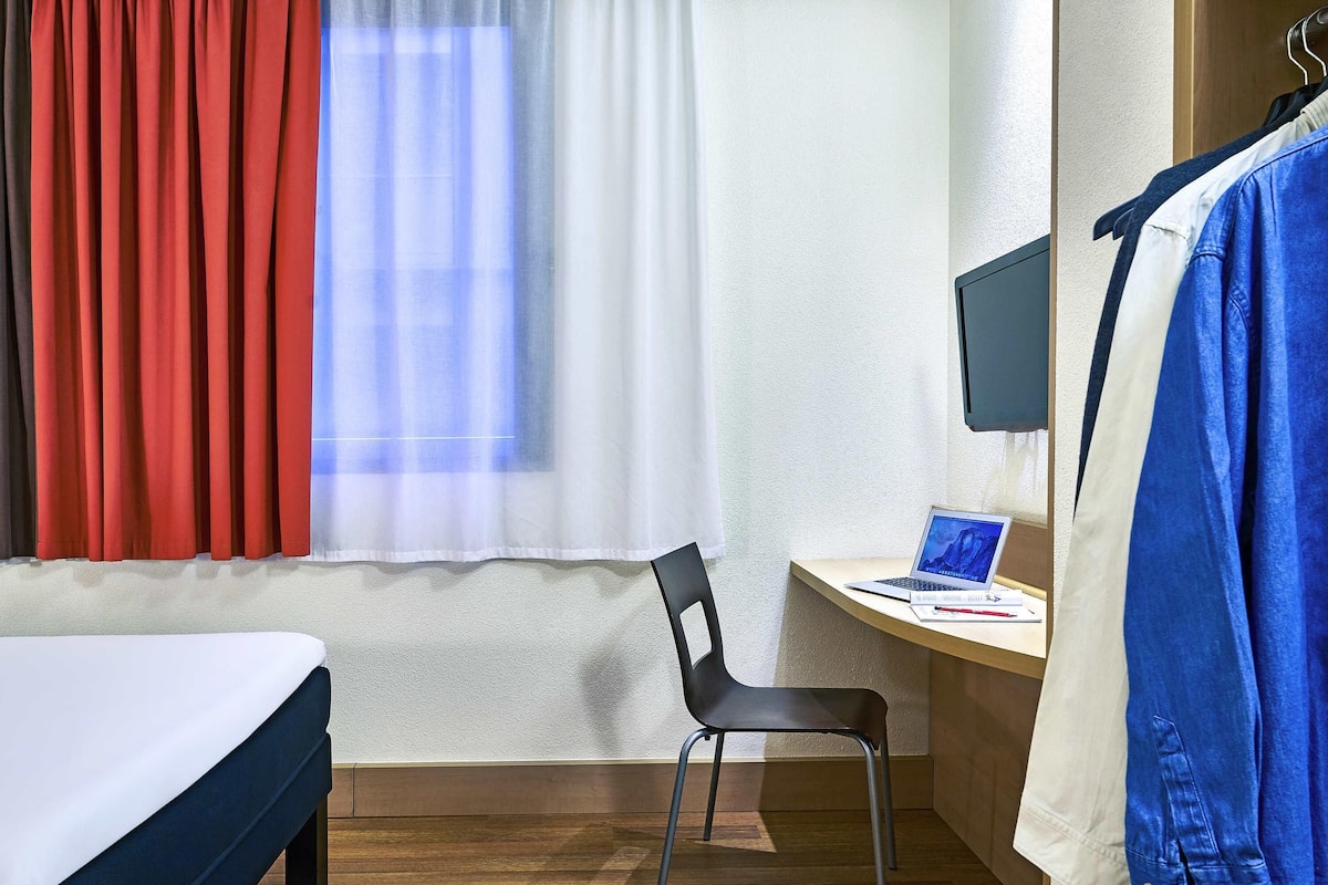 Habitación de hotel con cama, escritorio, laptop, silla y televisor en la pared.