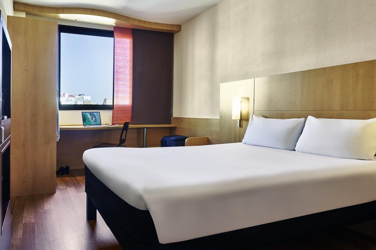 Une chambre d’hôtel avec un grand lit, un bureau avec un ordinateur portable, une fenêtre donnant sur un beau paysage et une lampe fixée au mur.