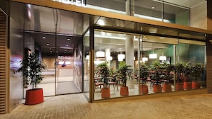 Une entrée d’hôtel moderne avec des portes vitrées, des plantes en pot et un espace restaurant bien visible à l’intérieur.