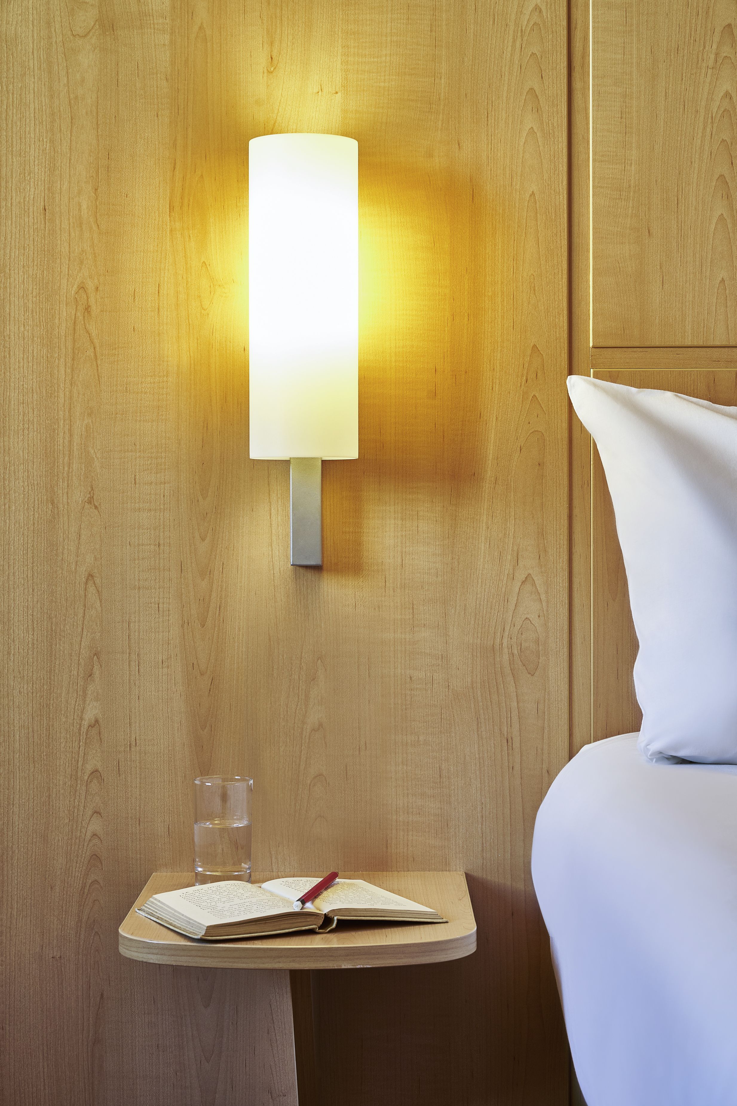 Un mur en bois, avec une lampe fixée au mur et une petite table de chevet sur laquelle se trouvent un livre et un verre d’eau.