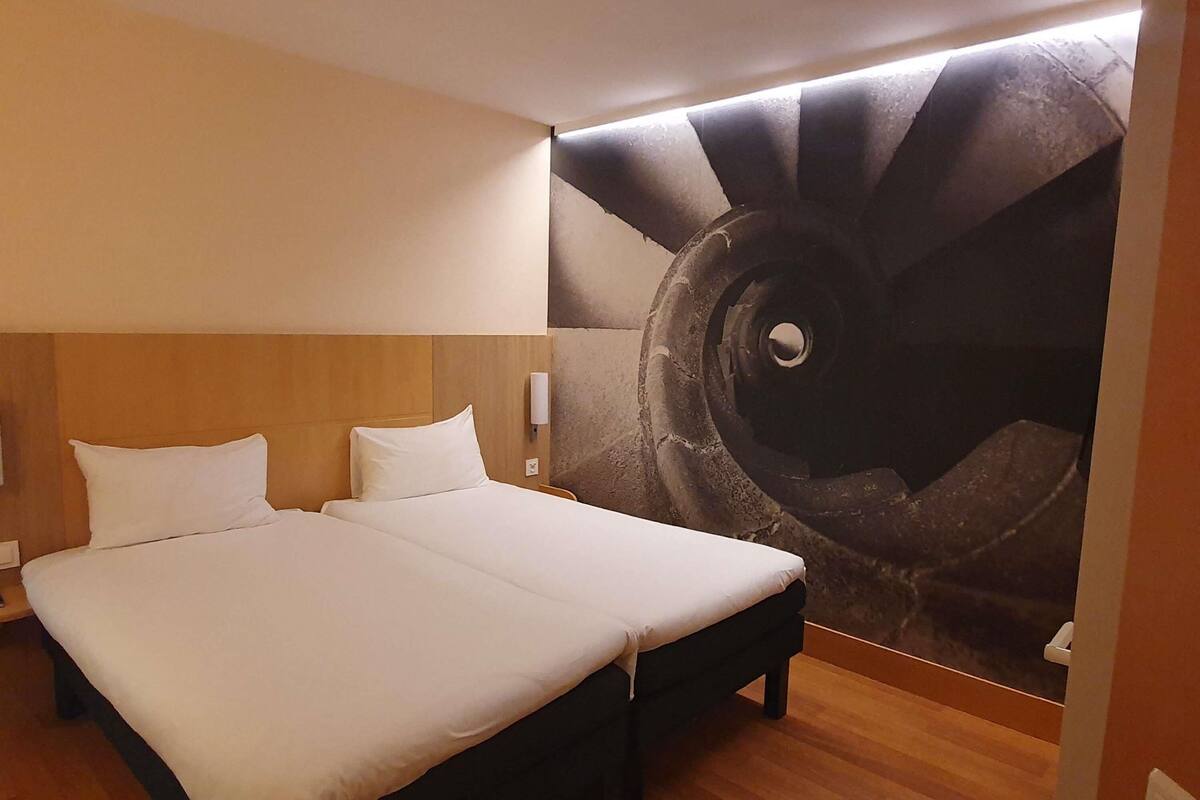 Habitación de hotel con una cama grande, cabecero, luz empotrada en la pared y un mural de una escalera de caracol.
