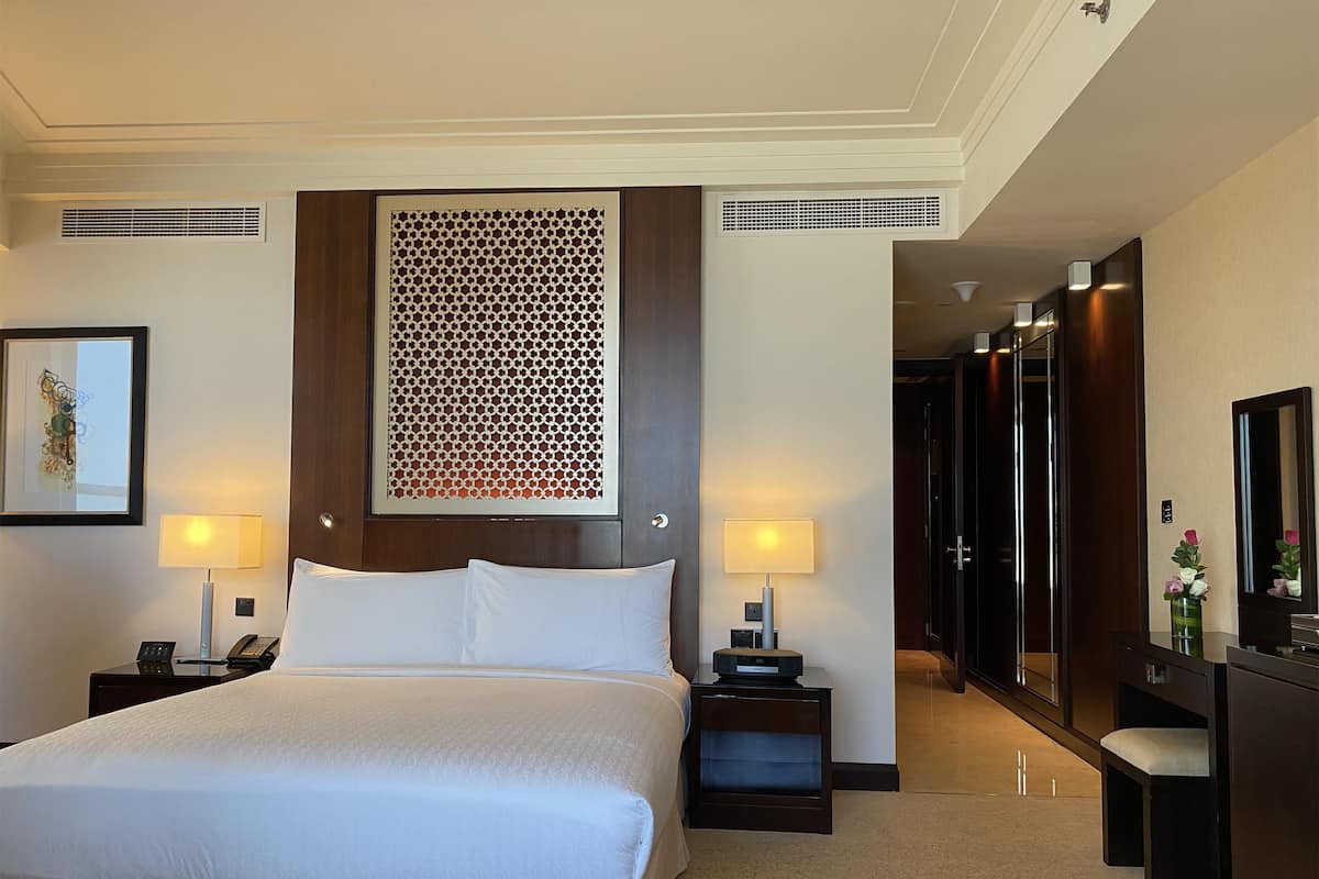 Une chambre d’hôtel avec un grand lit, deux tables de chevet avec des lampes, un panneau décoratif au mur, une œuvre d’art encadrée et un miroir.