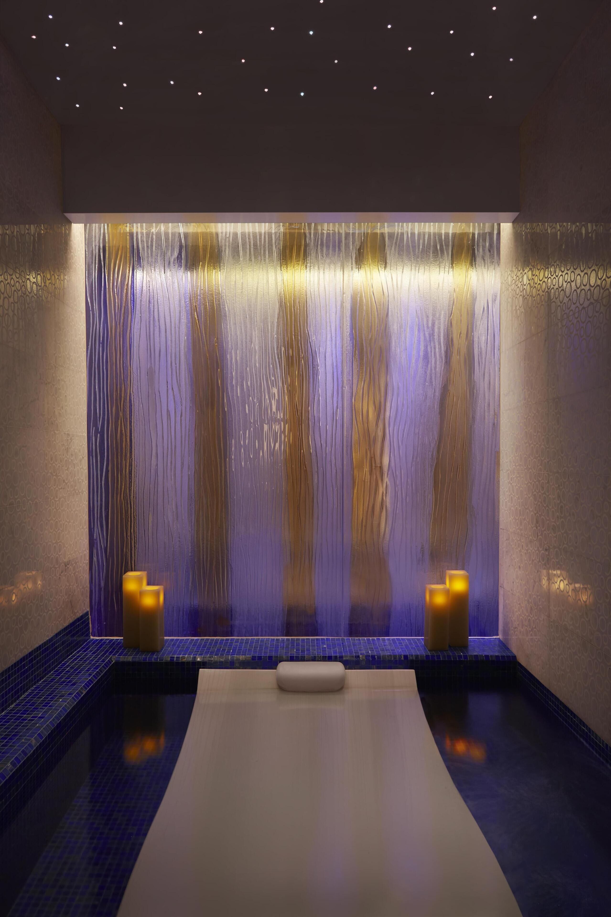 Un cuarto de spa con una gran fuente en la pared, una camilla de masaje y dos velas.