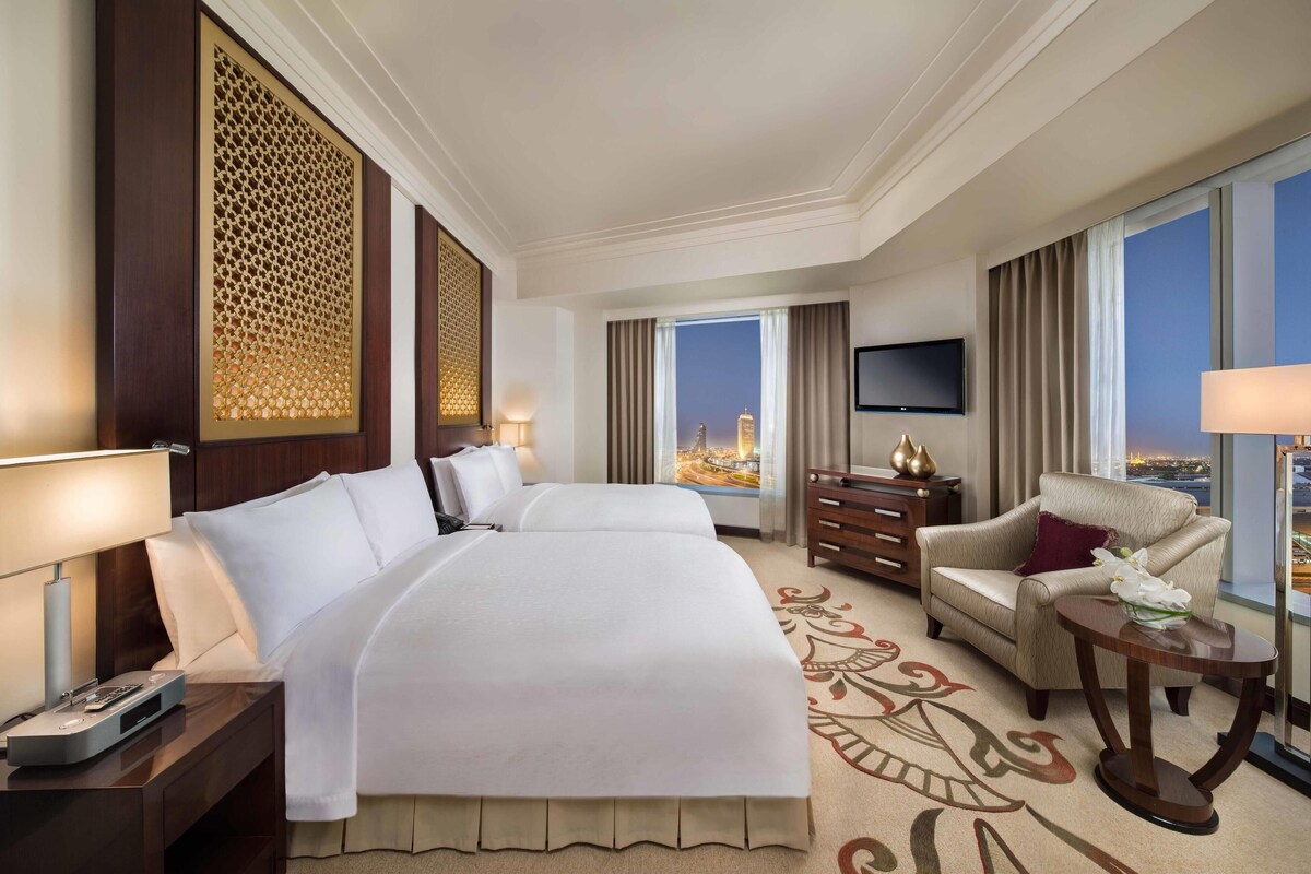 A hotel room with a large bed, a desk with a lamp, a chair, a TV on the wall, and a view of the cityscape.
