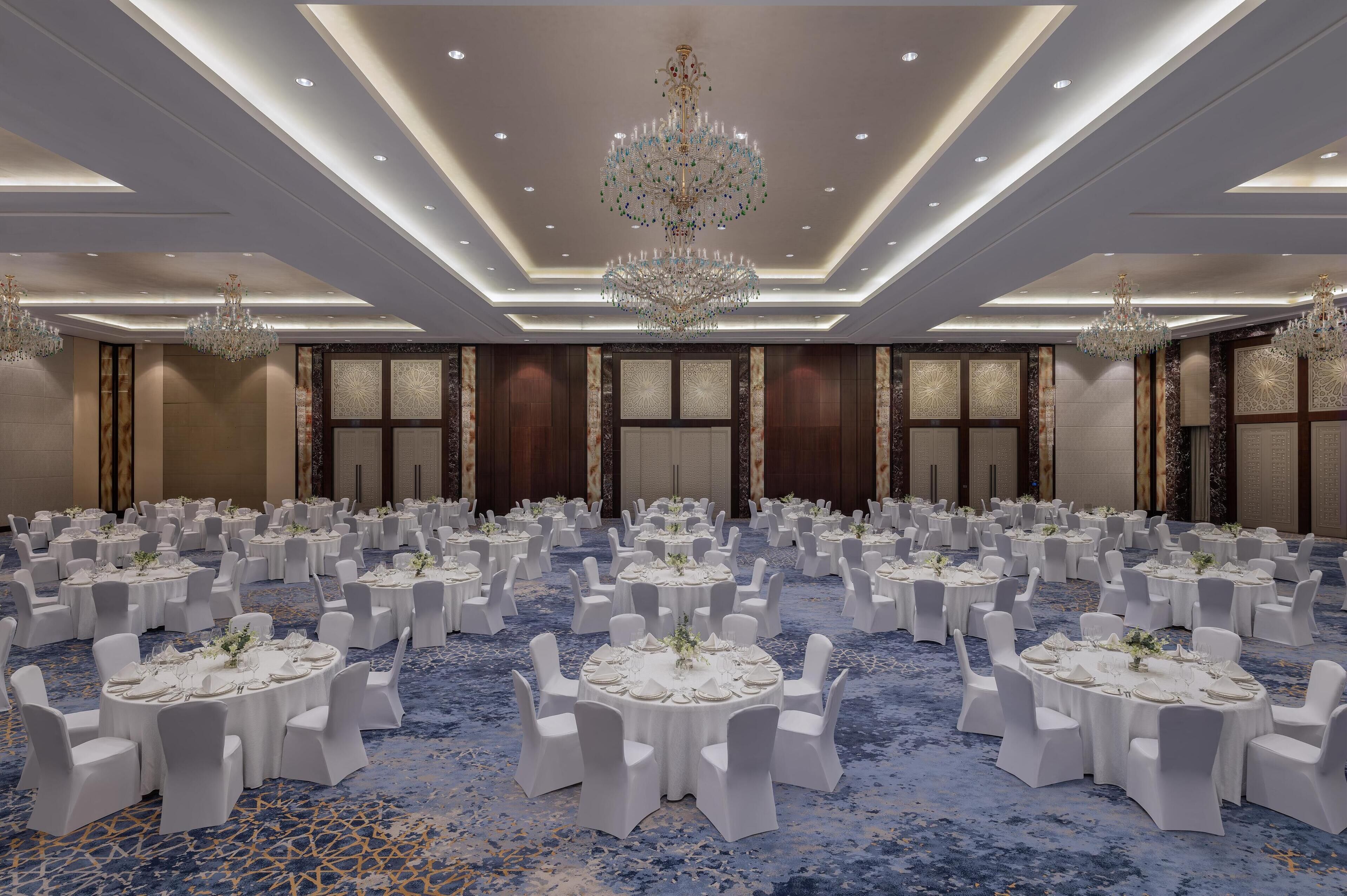 A banquet hall with round tables, white chairs, and chandeliers.