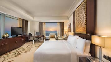 A hotel room with a large bed, a desk with a chair, a television, and a view of the sunset through the window.