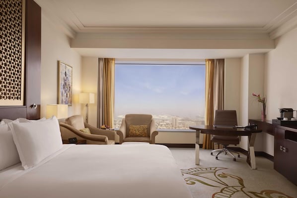 A hotel room with a large bed, two armchairs, a desk, and a view of a cityscape through large windows.