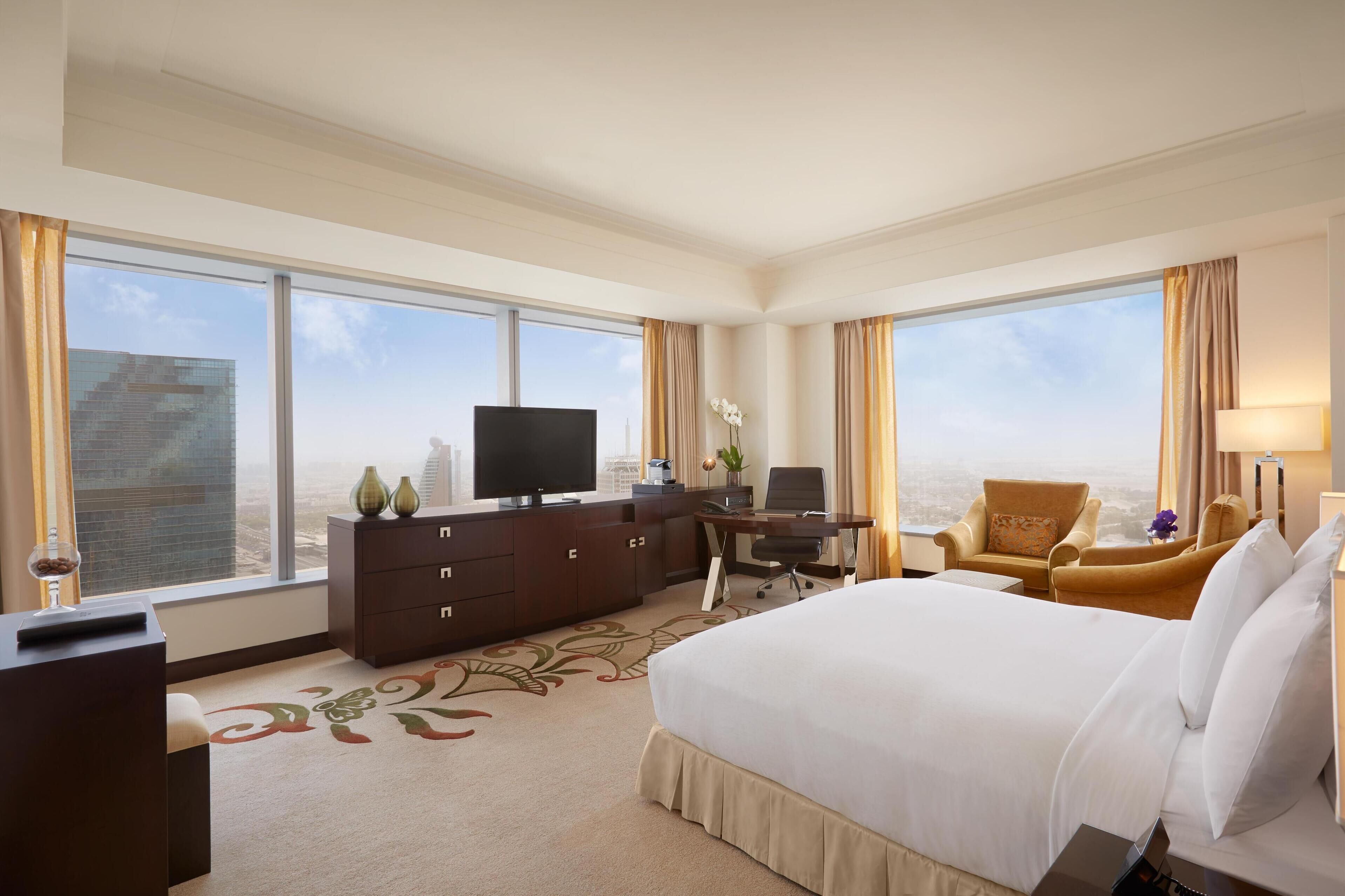 A hotel room with a large bed, a desk with a computer, a TV, and a view of the cityscape through large windows.