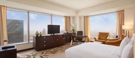 A hotel room with a large bed, a desk with a computer, a TV, and a view of the cityscape through large windows.
