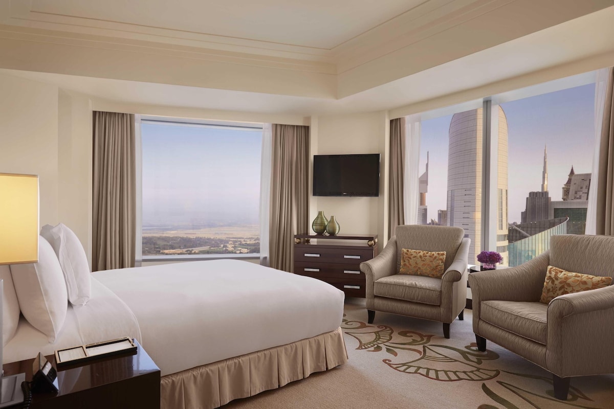 A hotel room with a large bed, two armchairs, a flat-screen TV, a nightstand, and a view of a cityscape.